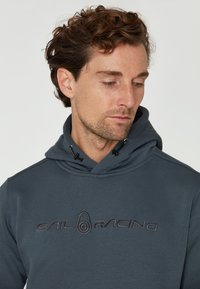 Mörkgrå hoodie med en stor broderad "SAIL RACING"-logotyp. Har en justerbar huva och textil med struktur. Närbild av plagget.
