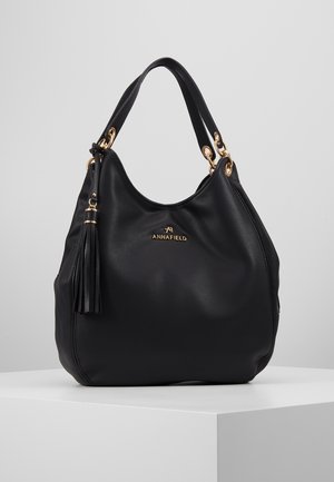 Schwarze Lederhandtasche mit goldenen Ringen und Quaste, geprägt mit dem "ANNAFIELD"-Logo, stehend auf weißer Oberfläche vor einem schlichten grauen Hintergrund.