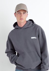 Sweat à capuche gris avec une poche avant, arborant un logo blanc sur la poitrine. Le mannequin porte également une casquette beige. Tissu lisse, coupe décontractée.