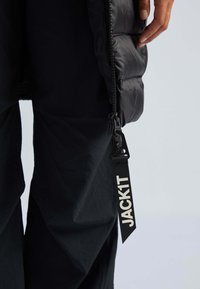 Giubbotto puffer nero con una superficie testurizzata. La chiusura con cerniera presenta una cintura in tessuto etichettata "JACK1T." Indossato sopra pantaloni scuri.