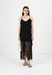 Robe noire avec des volants superposés, bretelles spaghetti réglables et section inférieure transparente. Associée à un petit sac blanc et des sandales à talons noires.