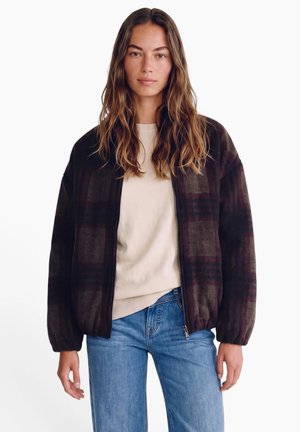 Femme aux longs cheveux ondulés porte une veste zippée à carreaux foncée par-dessus un pull beige et un jean bleu, debout sur un fond blanc.
