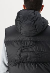 Chaleco de plumas negro con cuello alto y capucha, que presenta un logo de cocodrilo en relieve en la parte posterior y una textura suave y brillante.
