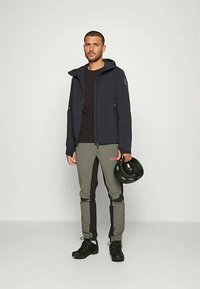 Férfi fekete dzsekit, fekete inget, szürke és fekete nadrágot, fekete cipőt visel, a jobb kezében egy fekete sisakot tart, és egy sima fal előtt áll.