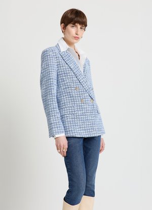 Blazer doppiopetto in tweed azzurro chiaro con colletto, bottoni dorati e motivo a trama, abbinato a jeans scuri e stivali al ginocchio.