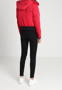 Červená prošívaná bunda s kapucí a žebrovaným černým lemem, kombinovaná s černými skinny džíny a bílými teniskami, zobrazená zezadu.