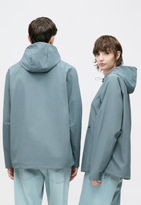 Lyseblå hætte-regnjakke med afslappet pasform, der har sidelommer og en snøre i hætten. Båret sammen med lyseblå denim jeans.