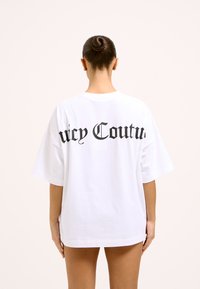 Valge, ülimõõdus puuvillane t-särk, mille tagaküljel on must "Juicy Couture" tekst, ümarnööriga kaelus ja lühikesed varrukad.