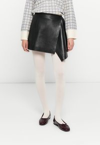 Donna che indossa una mini gonna in pelle nera asimmetrica, collant bianchi, ballerine bordeaux con fiocchi e un maglione a maniche lunghe a fantasia.