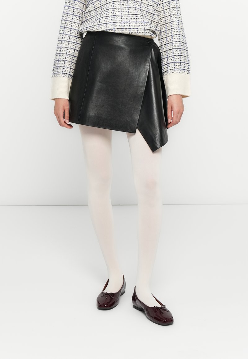Donna che indossa una mini gonna in pelle nera asimmetrica, collant bianchi, ballerine bordeaux con fiocchi e un maglione a maniche lunghe a fantasia.