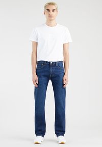 Jauns cilvēks ar īsiem blondiem matiem, tērpies baltā T-kreklā, tumši zilos taisnos džinsos un baltās kedās, stāv uz vienkārša fona.