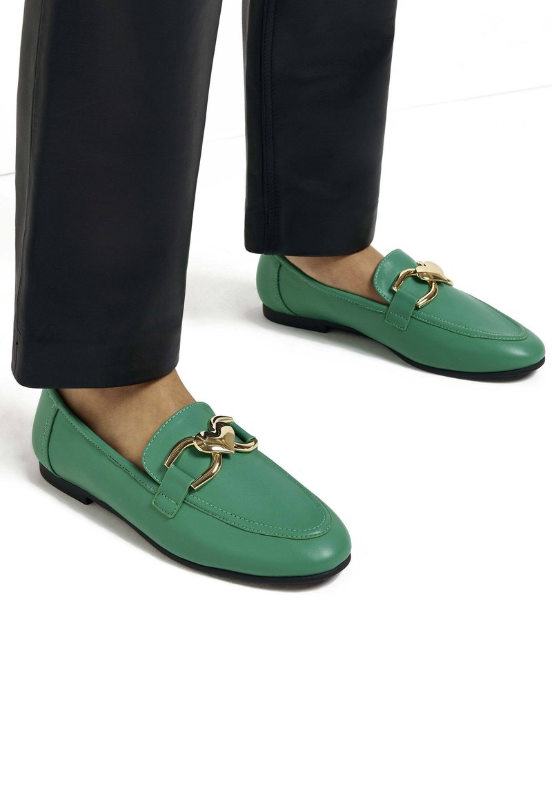 Personne portant des mocassins en cuir verts avec un détail en métal doré en forme de cœur et un pantalon noir sur fond blanc.