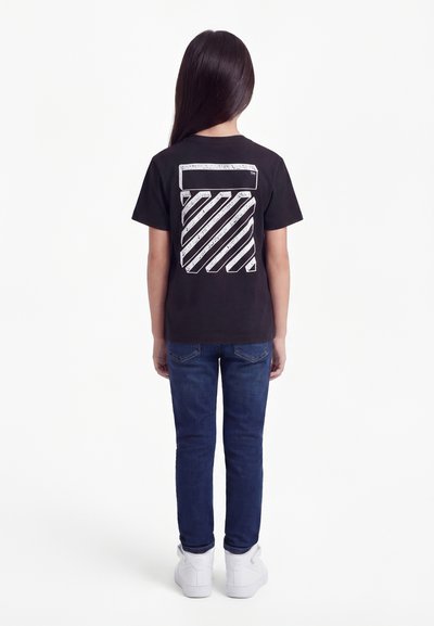 Παιδί με μακριά σκούρα μαλλιά, φοράει μαύρο μπλουζάκι με σχέδιο, μπλε τζιν και λευκά αθλητικά παπούτσια, στέκεται με την πλάτη γυρισμένη σε απλό λευκό φόντο.