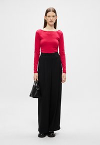 Rdeča bluza z dolgimi rokavi, črne široke hlače z visokim pasom, črna torbica in debela črna obutev. Enostaven dizajn, gladka tekstura tkanine.