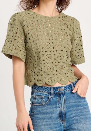 Crop top en dentelle crochet vert olive avec manches courtes et ourlet festonné, porté avec un jean en denim bleu taille haute.
