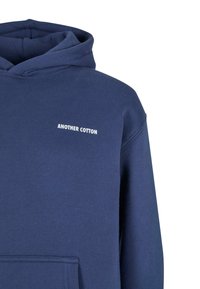 Granatowy hoodie wykonany z bawełny; ma przednią kieszeń i biały napis logo "ANOTHER COTTON" na piersi. Gładka tekstura.
