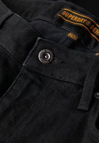 Czarne dżinsy mają wyraźne zapięcie na guzik z wygrawerowanym logo. Tkanina jest gładka w dotyku, a szwy są widoczne wzdłuż krawędzi.