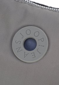 Šedý látkový povrch s kulatou šedou gumovou nášivkou s logem "JOOP! JEANS" kolem středního otvoru.