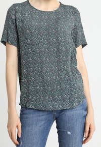 Kvinde iført en mørkegrøn kortærmet bluse med små hvide og teal blomsterprint, parret med blå slidte jeans.