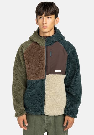 Jongeman in een multicolor patchwork fleece jack met rits aan de voorkant en capuchon, staande tegen een plain achtergrond.