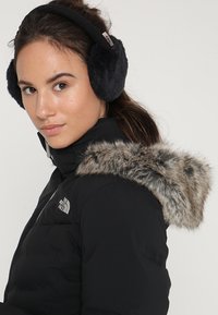 Chaqueta puffer negra con capucha de piel sintética desmontable, presentada con un logo plateado en el pecho. Modelo llevando orejeras de terciopelo negras.