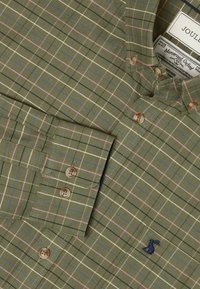 Chemise à carreaux verts en tissu à motifs, avec des rayures bleu clair, rose et blanc. Détails avec des boutons marron et un logo brodé.