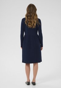 Femme aux cheveux ondulés portant une robe midi plissée bleu marine et des chaussures slip-on noires à clous, debout de dos sur un fond uni.