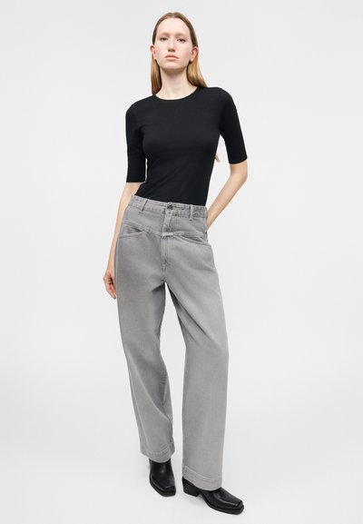 Γυναίκα που στέκεται με το χέρι στην τσέπη φορώντας μαύρο εφαρμοστό τοπ με μανίκια τρία τέταρτα, φαρδιά γκρι τζιν και μαύρα μποτάκια αστραγάλου σε απλό φόντο.