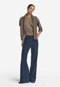 Maglione gilet marrone sopra una maglia taupe, abbinato a jeans scuri a gamba larga. Il modello indossa scarpe marroni e orecchini d'argento, in posizione eretta.
