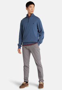 Maglione blu con zip a un quarto e polsini a coste. Indossato sopra una camicia a collo bordeaux, abbinato a pantaloni grigi e scarpe marroni.