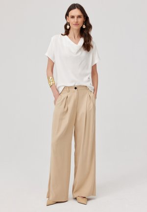 Kobieta nosząca białą bluzkę z krótkim rękawem, beżowe szerokie spodnie, beżowe buty w szpic, złotą bransoletkę i duże kolczyki, stojąca z rękami w kieszeniach.