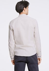Camisa de manga larga beige claro con cuello, tejido texturizado y puños abotonados. Presenta un dobladillo curvado y un solo yugo trasero.