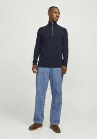 Marineblauer Ripp-Pullover mit hohem Kragen und silbernem Reißverschluss, kombiniert mit hellblauen weiten Jeans und dunkelbraunen Schuhen.