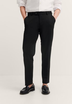 Mann trägt schlanke schwarze Anzughose, weißes Hemd mit hochgekrempelten Ärmeln, schwarze Leder-Loafer, steht mit Händen in den Taschen.