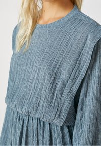 Mujer vistiendo un vestido largo de manga larga, texturizado y azul, con un amplio cuello cuadrado y cintura fruncida, mostrado desde los hombros hasta la mitad del torso.