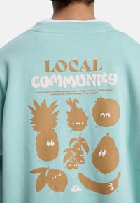 Πολυτελές φούτερ μέντας με γραφικό τύπωμα καρτούν φρούτων σε χρυσό και λευκό, με παιχνιδιάρικα μάτια και το κείμενο «ΤΟΠΙΚΗ ΚΟΙΝΟΤΗΤΑ» επάνω.