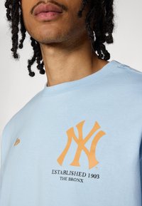 T-shirt en coton bleu clair avec un logo orange des New York Yankees, accompagné de "ÉTABLI EN 1903 - LE BRONX" en noir sous le design.