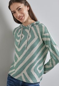 Hoodie vert clair avec un motif abstrait blanc, cordon de serrage et tissu texturé. Stylisé avec des manches retroussées et associé à un jean bleu.