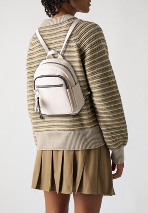 Persoon die een gestreepte gebreide trui en een beige geplooide rok draagt, met een kleine witte leren rugzak op de rug, van achteren gezien.