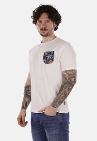 Camiseta de algodón blanca con un bolsillo en el pecho que presenta un estampado tropical de palmeras y flores. Diseño de manga corta y cuello redondo.