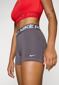 Szare szorty Nike Pro z szerokim czarnym pasem w talii, ozdobione białym napisem, gładką fakturą i małym białym logo po boku.