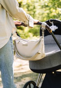 Lichtbeige schoudertas gemaakt van zacht materiaal, met een ritssluiting en verstelbare riem, bevestigd aan een kinderwagen met een donkergrijze zitplaats.
