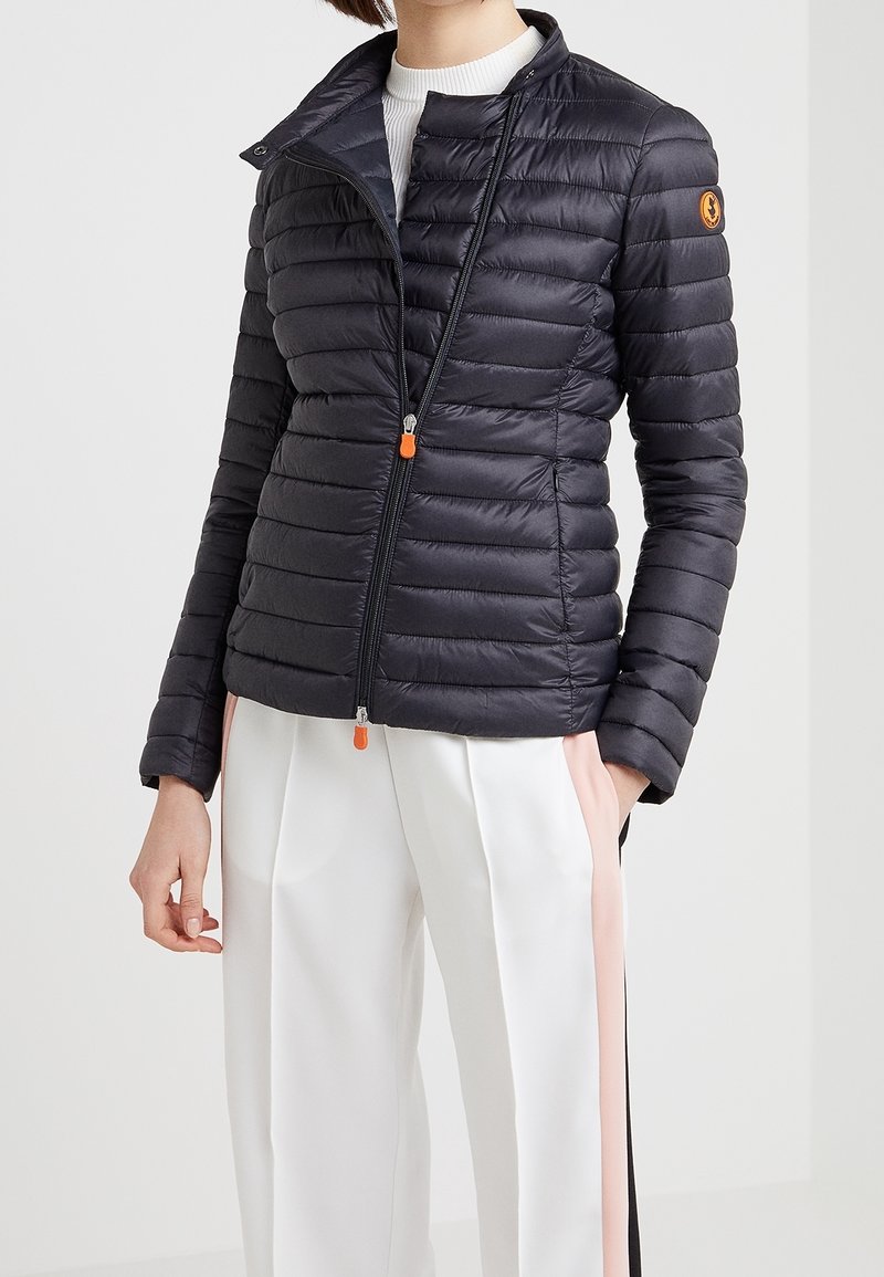 Femme portant une veste matelassée noire avec des tirettes de fermeture éclair orange et un pantalon blanc avec des bandes latérales roses et noires, debout devant un fond uni.