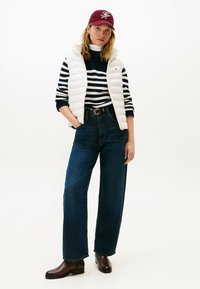 Gilet imbottito bianco indossato sopra un maglione a righe blu navy e bianco, jeans a gamba larga blu scuro e stivaletti marroni; berretto bordeaux con logo.