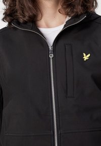 Zwarte jas gemaakt van zachte stof, voorzien van een zilveren rits, een voorzak en een klein geel vogellogo op de linkerborst.