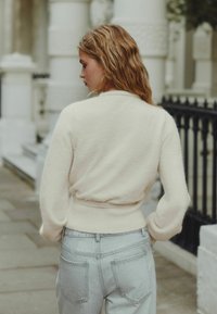 Cream knitted sweater with a fitted waist, cozy texture. Paired with light-wash jeans, featuring back pockets and a relaxed fit.