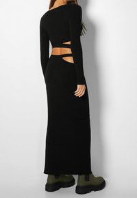 Czarna sukienka z długim rękawem w prążki, z wycięciami w talii, o dopasowanym kroju, uzupełniona zielonymi botkami o długości do kostek.