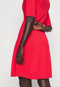 Robe en maille rouge à manches courtes, texture côtelée, avec des détails de couture décorative sur les côtés. Silhouette trapèze avec un corsage ajusté.