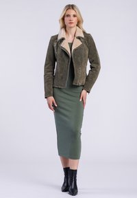 Model in een olijfgroen suede jack met een crèmekleurige kraag, gecombineerd met een getailleerde groene jurk en zwarte enkellaarsjes, staat zelfverzekerd.