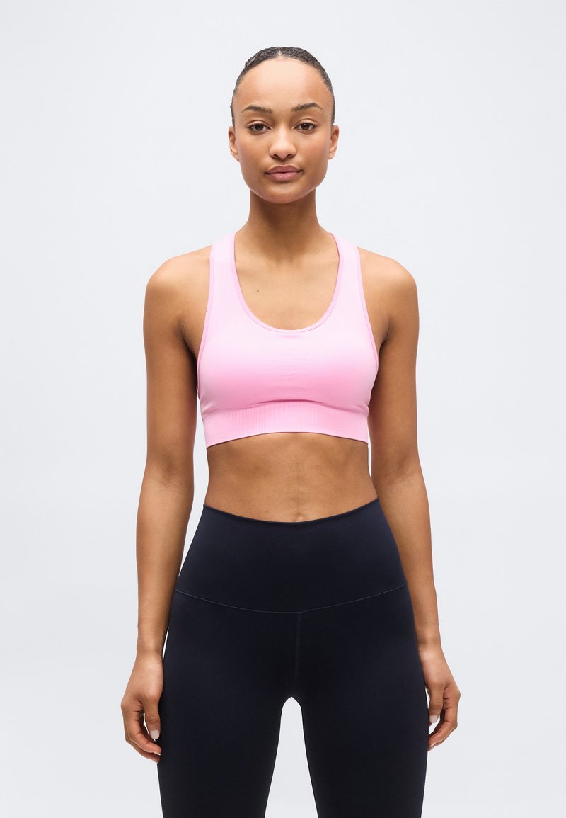 Νεαρή γυναίκα που στέκεται, φορώντας ένα ανοιχτό ροζ αθλητικό τοπ και μαύρο ψηλόμεσο κολάν, μπροστά σε ένα απλό ανοιχτό φόντο.